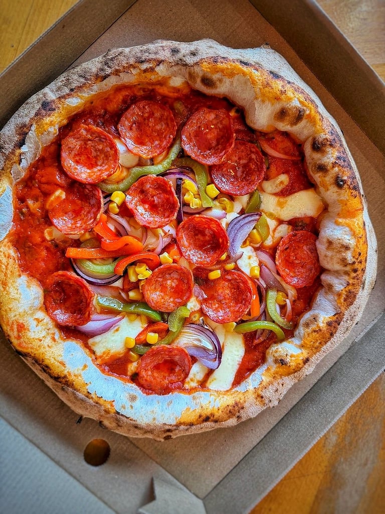 Wood-fired pizza with pepperoni, corn, red onions, and green peppers on charred crust in cardboard box