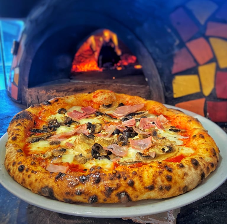 Wood-fired pizza with ham, olives, and cheese on white plate with traditional brick oven in background