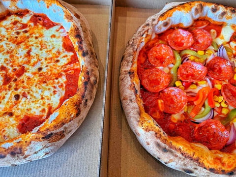 Two wood-fired pizzas with melted cheese and red toppings, displayed in cardboard boxes on a wooden surface