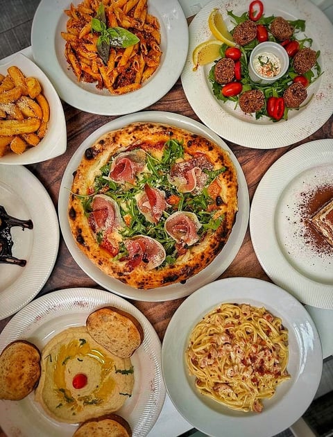 Overhead view of Italian restaurant dishes including pizza with prosciutto, pasta carbonara, meatballs, bruschetta, and fried potatoes on white plates