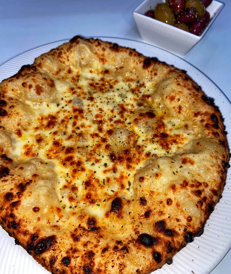 Golden-brown pizza with melted cheese and spices on white plate, small bowl of olives in background