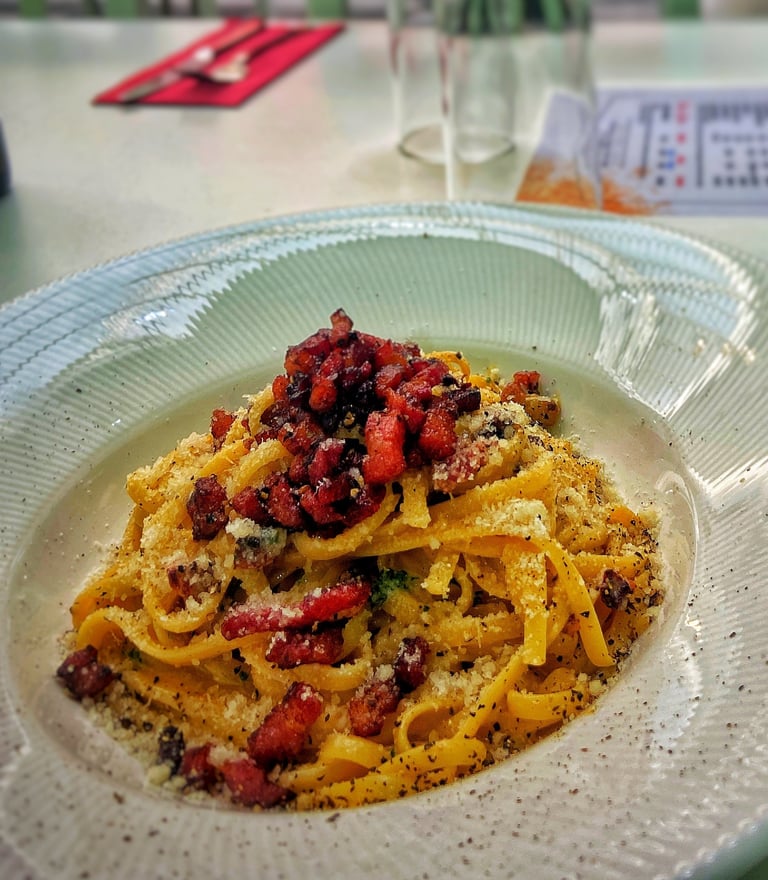 Bowl of golden pasta with red peppers and black crumble topping served on a textured white plate at a dining table
