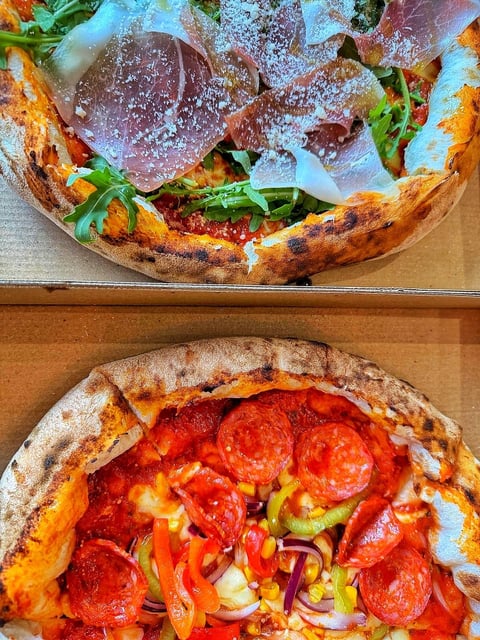 Two wood-fired pizzas stacked, top one with prosciutto and arugula, bottom one with pepperoni and bell peppers