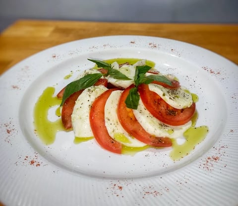 Caprese salad with alternating tomato and mozzarella slices, fresh basil, olive oil drizzle, and red pepper seasoning on a white plate.