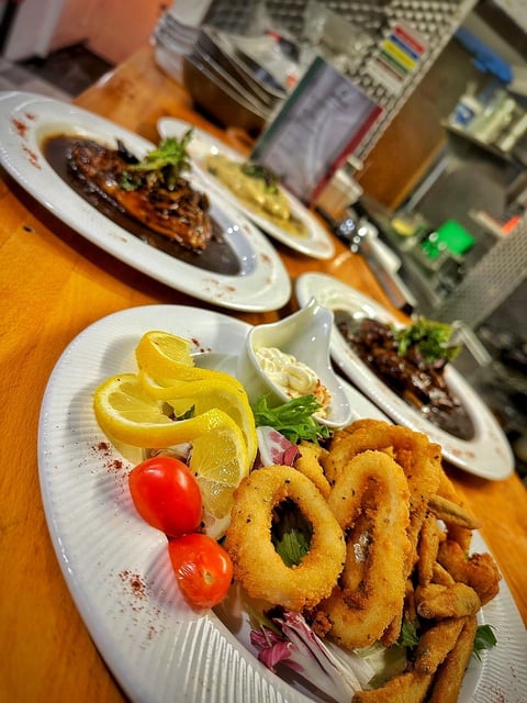 Restaurant plates with breaded calamari rings, lemon slices, cherry tomatoes, and grilled meat dishes on wooden table