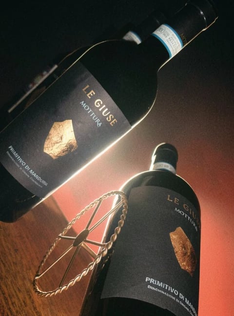 Two dark wine bottles with gold leaf labels displayed at an angle on a warm-toned background with rope accent
