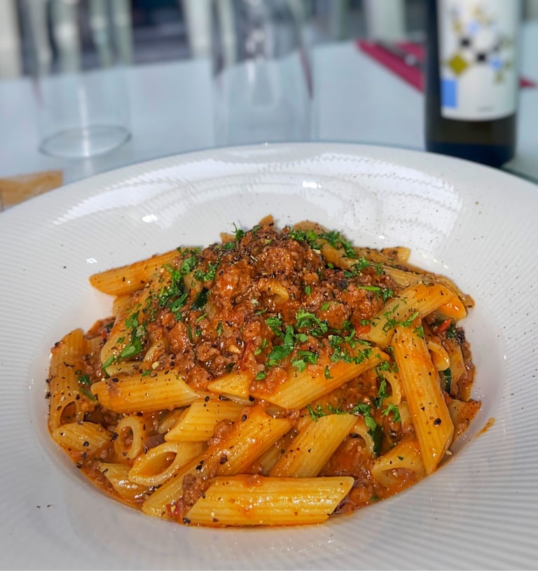 Plate of penne pasta with meat sauce and fresh parsley garnish in a restaurant setting with wine bottle and glasses in background