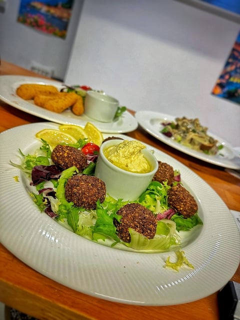 Plate of fried falafels on mixed greens with lemon slices and yogurt sauce, served with additional dishes in background